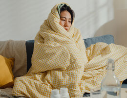 A woman wrapped in a blanket on a couch with water and pill bottles on a coffee table.