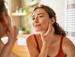 A woman's reflection in a mirror as she smiles and touches her face.
