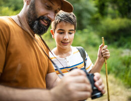 A man and boy on a hiking trail look at the man’s binoculars.